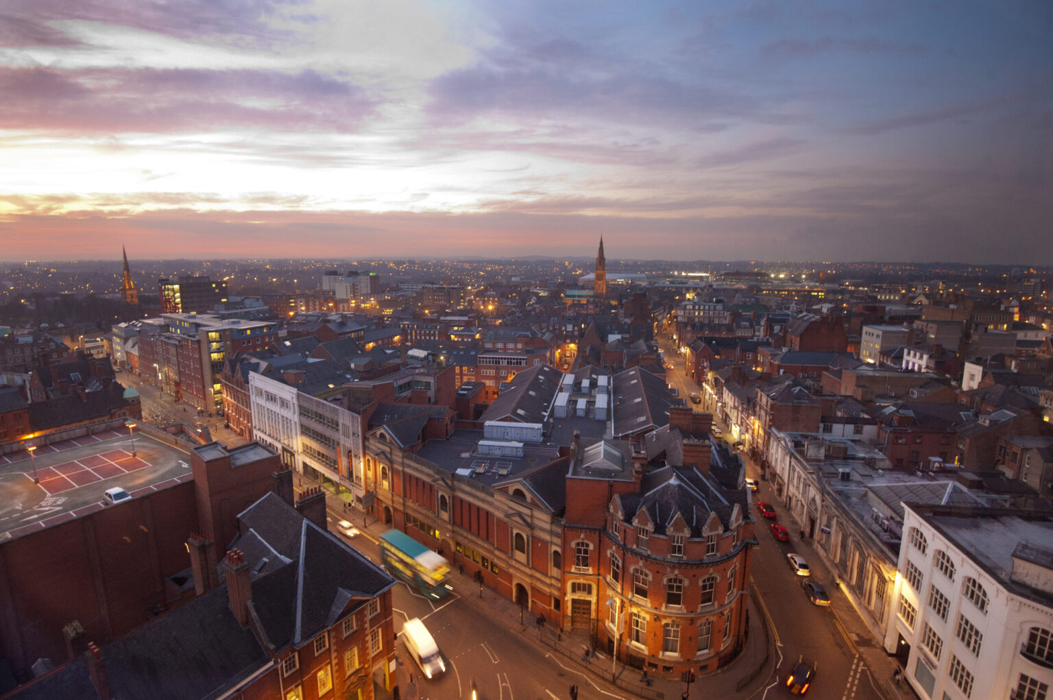Leicester city skyline at dusk with Leicester Cathedral spire and red-brick rooftops under a pink sunset sky